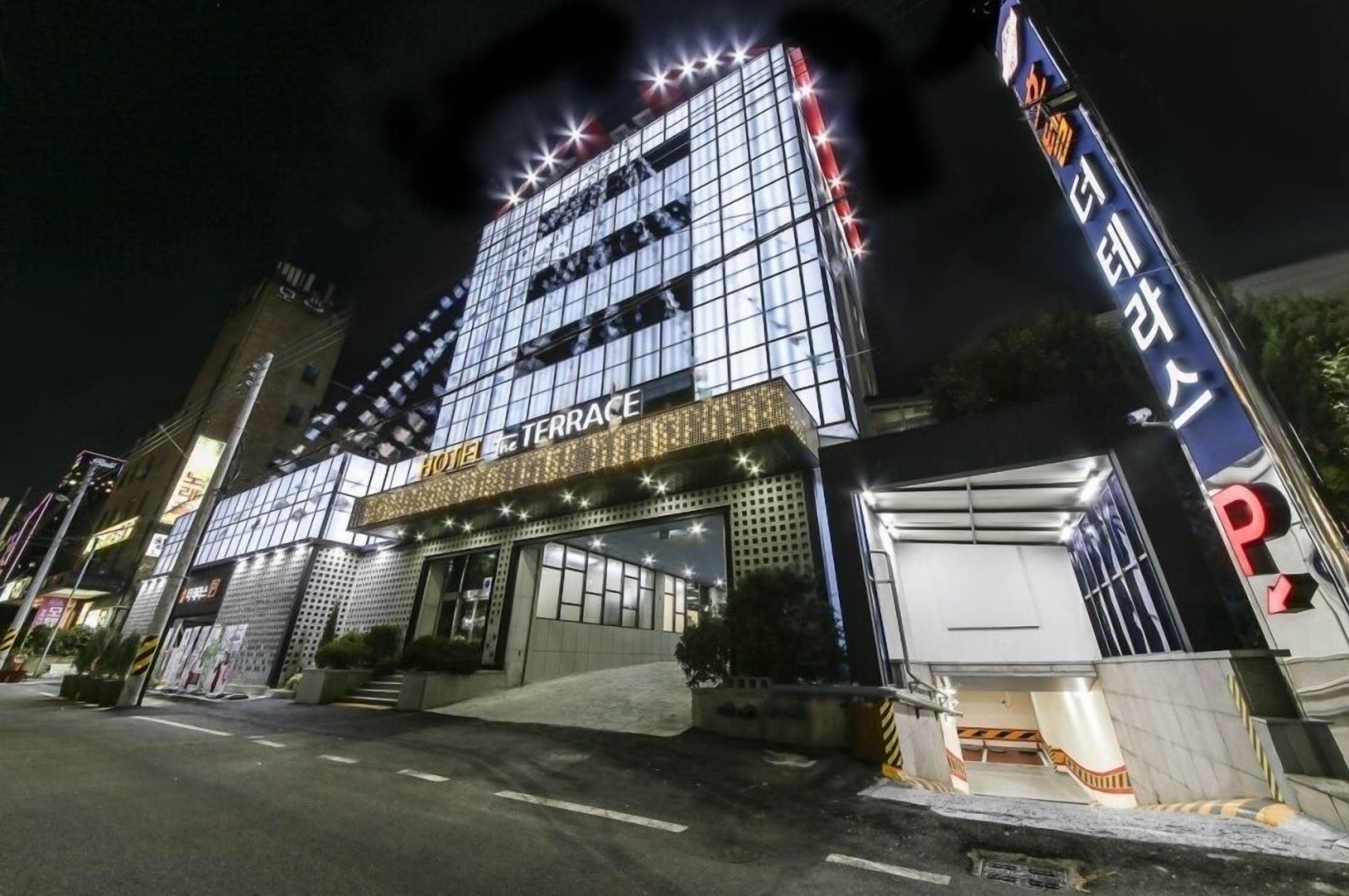 A modern hotel with a glass facade, a prominent terrace, and a parking area.