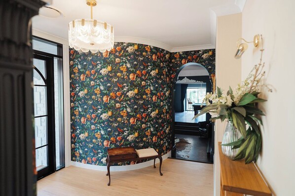 A room with a floral wallpaper, a bench, a chandelier, and a vase with flowers.