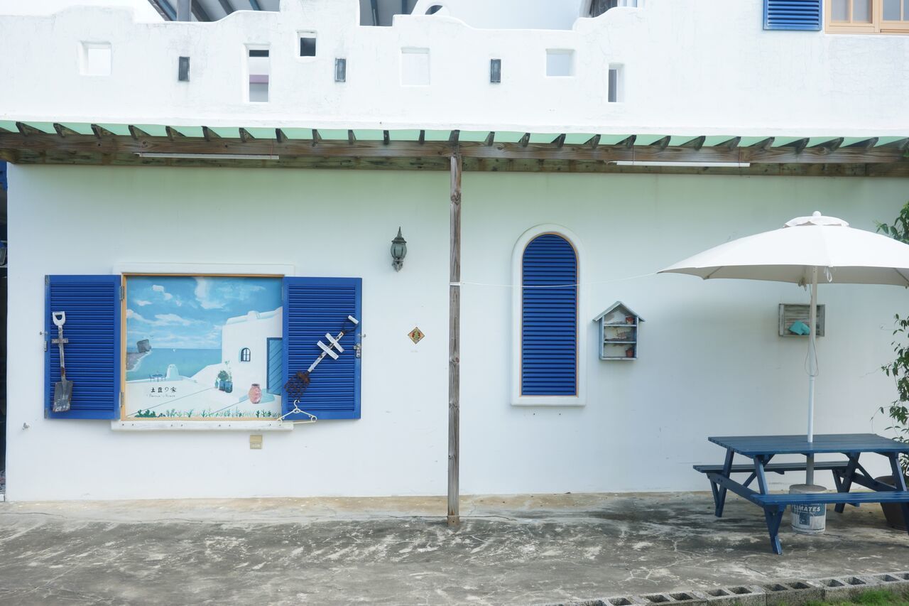 A white building with blue shutters, a wooden table with a white umbrella, and a poster of a coastal scene.