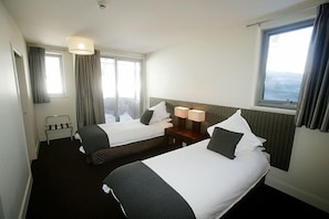 Habitación de hotel con dos camas, una mesita con lámparas, un ventanal con cortinas y vistas al exterior.
