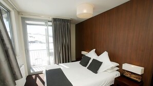A hotel room with a large bed, a bedside table with a lamp, a view of the outdoors through a window, and a wooden wall.