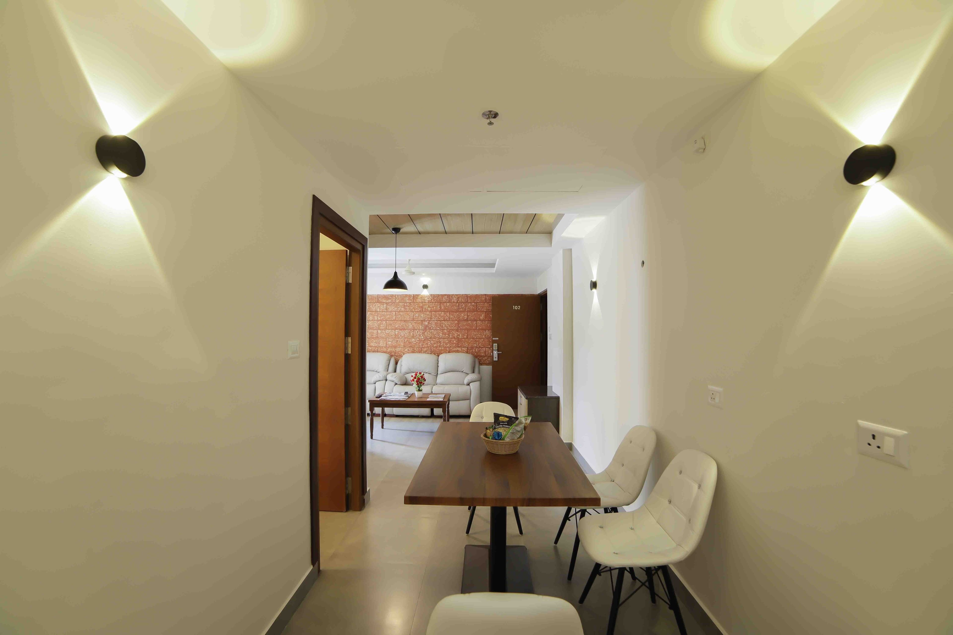 Un espace repas moderne avec une table en bois et des chaises blanches, un mur en briques en arrière-plan et un éclairage encastré au plafond.