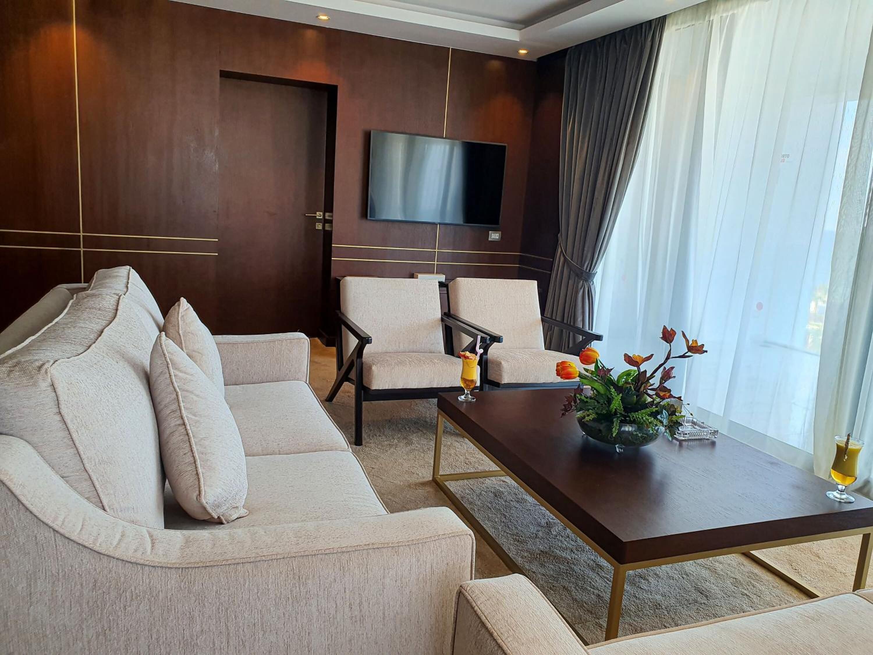 A modern hotel room with a beige sofa, two armchairs, a coffee table with a vase of flowers, and a flat-screen TV mounted on the wall.