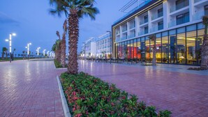 A modern hotel with a glass facade, outdoor seating area, and palm trees. - Porto Said Hotel & Spa (Al-Manasrah)