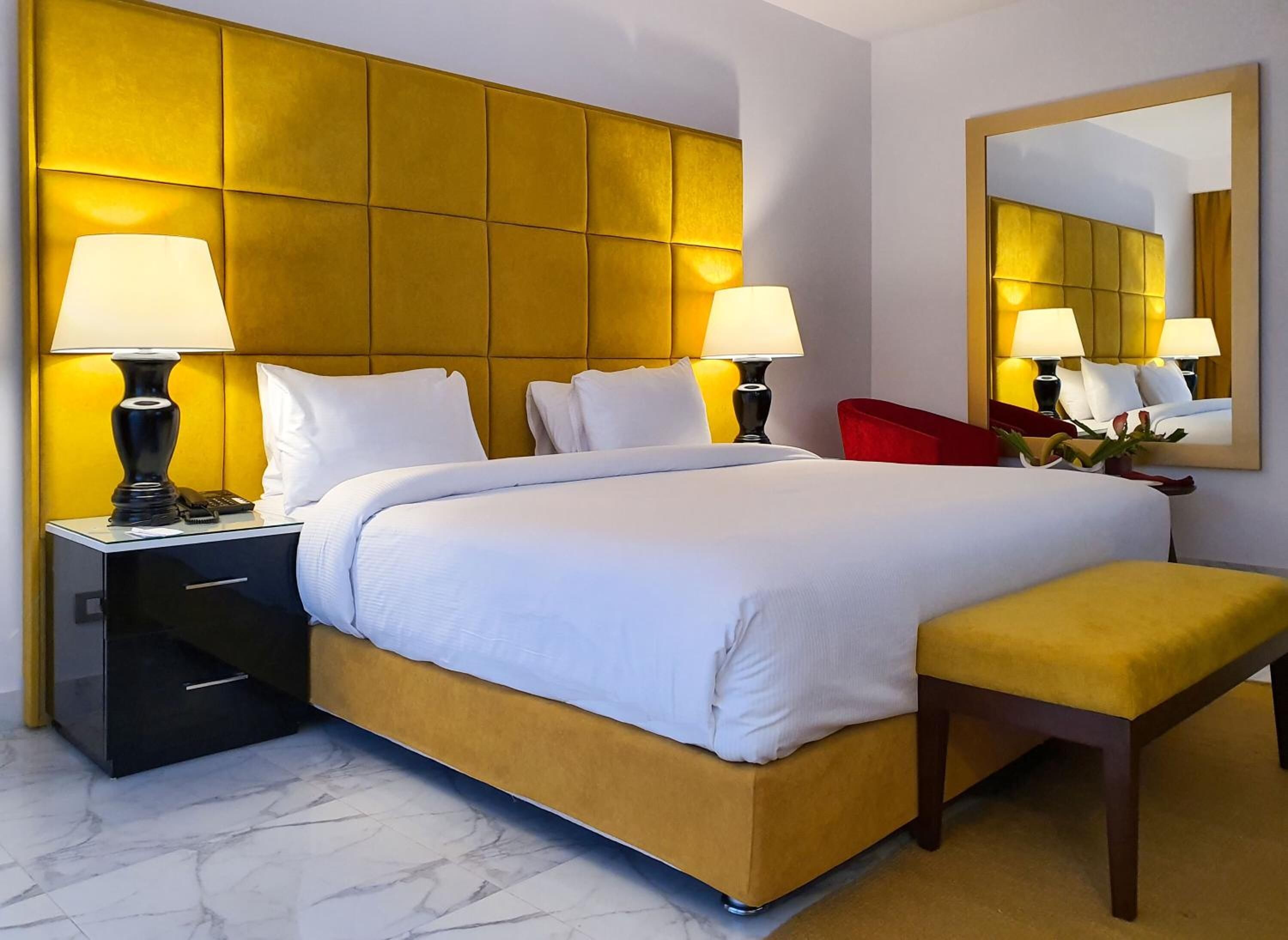 Ein Hotelzimmer mit einem großen Bett, zwei Nachttischen mit Lampen, einem roten Sessel, einer Bank und einem großen Spiegel an der Wand.