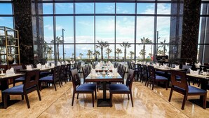 A restaurant with a long table set for a meal, large windows, and a view of palm trees and the ocean. - Porto Said Hotel & Spa (Al-Manasrah)