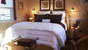 A neatly made bed with white bedding, two framed pictures on the wall above it, a bedside lamp, and a chair to the right.