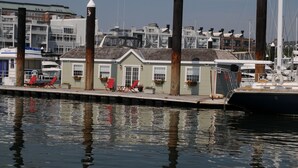 A marina with docked boats, a small building with red chairs, and a row of houses in the background.