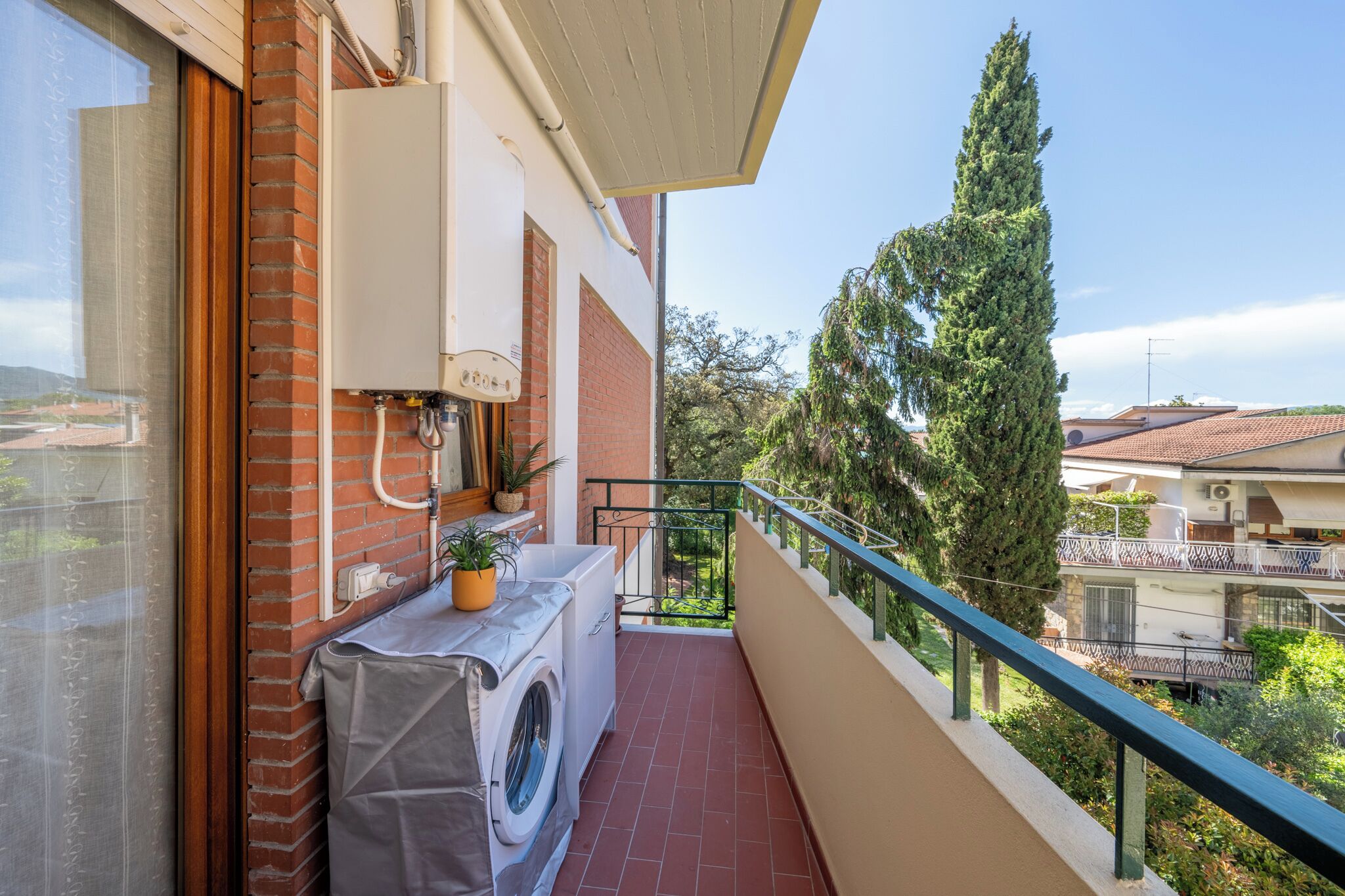 Een balkon met een wasmachine, een bloempot en uitzicht op omliggende huizen en bomen.