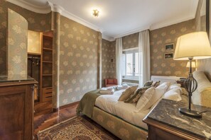 A bedroom with a bed, a nightstand, a lamp, a window, a radiator, a wardrobe, and a door.