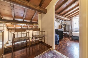 Quarto com piso de madeira, um banco com almofadas, uma escrivaninha e uma cama.