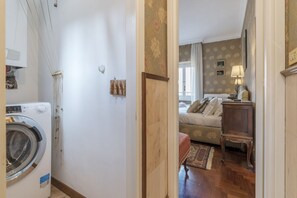 Uma área de lavanderia com máquina de lavar, uma porta de madeira que dá para um quarto com cama, mesa de cabeceira e abajur.