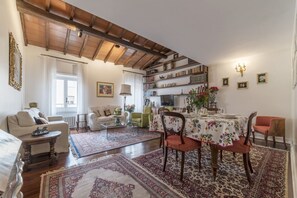 A spacious living room with a high ceiling, a dining area with a floral tablecloth, and a bookshelf filled with books.