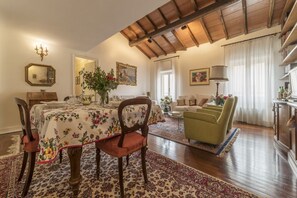 Uma sala de estar ampla com teto de madeira, um espaço de refeições com toalha de mesa florida e uma poltrona verde.