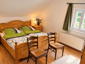 Ein Schlafzimmer mit einem hölzernen Bett, zwei Stühlen, einem Nachttisch mit Lampe, einem Fenster mit Vorhängen und einer Heizung.