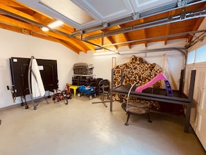 Eine Garage mit Betonboden, Holzträgern, einem Deckenventilator, einem Tisch mit einem rosa Stuhl, einem Holzstapel und verschiedenen Gegenständen, darunter ein Kinderwagen und ein Fahrrad.
