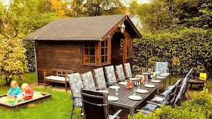 A wooden cabin with a dining table set for six, surrounded by chairs, in a lush garden.