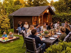 An outdoor dining area with a wooden structure, surrounded by greenery, and people seated at a table with food and drinks.