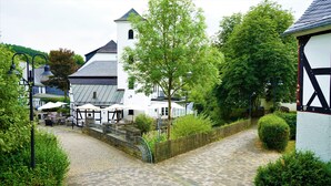 A cobblestone pathway leading to a white building with a black and white facade, surrounded by greenery and a small bridge.