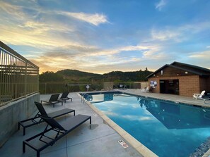 Een zwembad met ligbedden en een klein gebouw, omringd door een hek en heuvels op de achtergrond.
