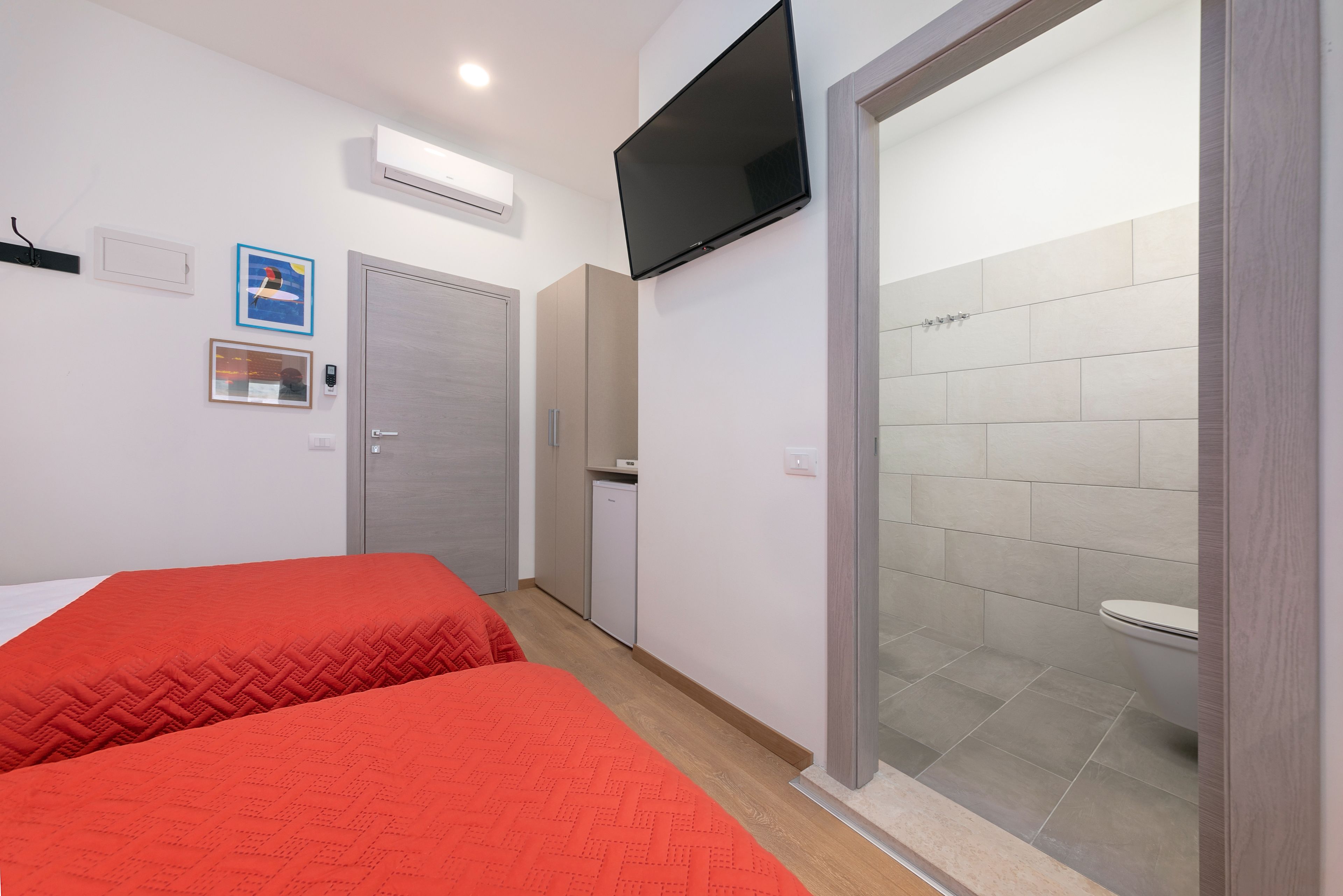A hotel room with two beds, a TV mounted on the wall, a small kitchenette, and a bathroom with a toilet and shower.
