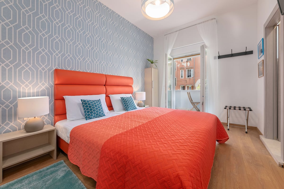 A modern bedroom with a geometric patterned wall, a bright orange headboard, and a view of buildings through a window.