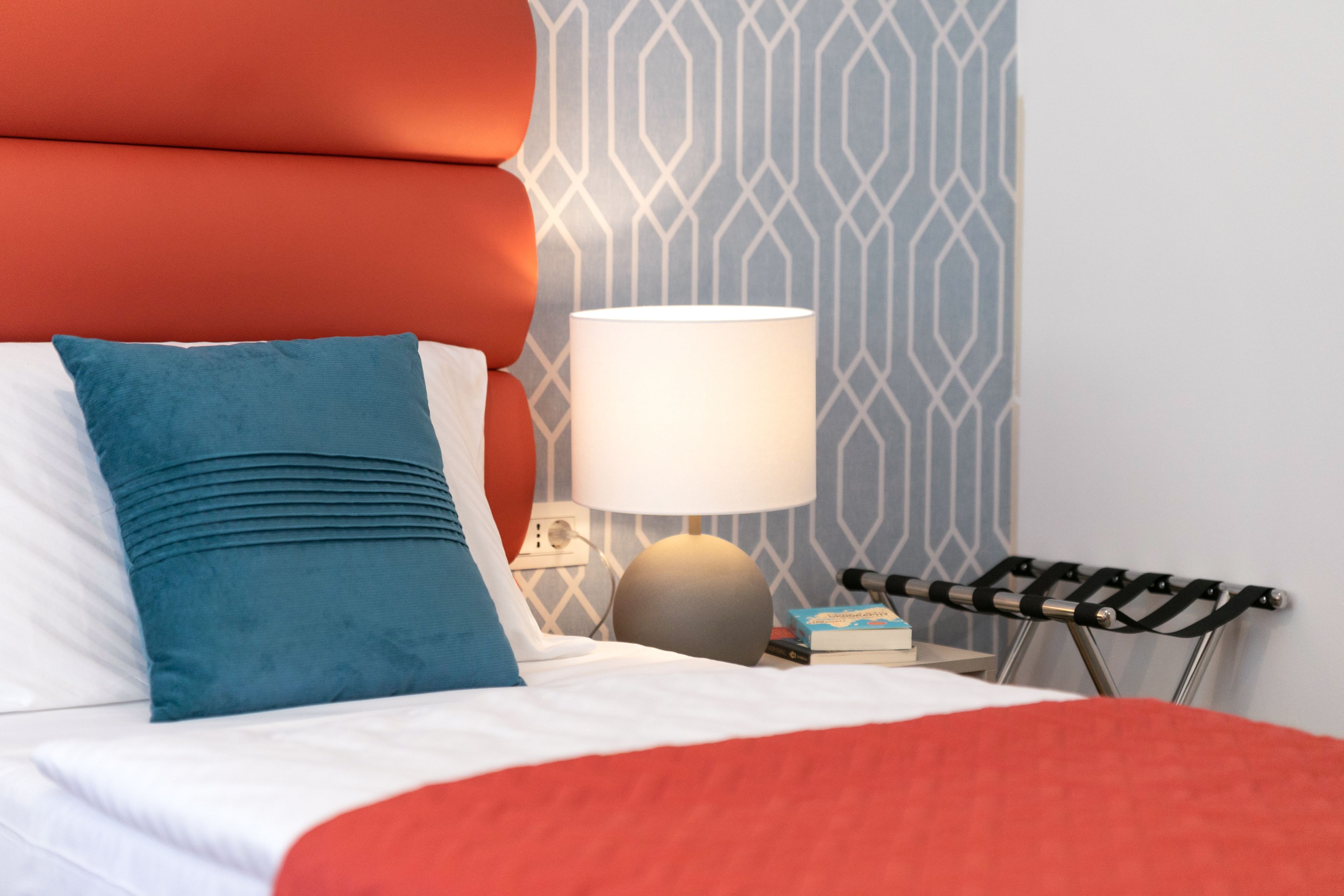 A neatly made bed with a red blanket, a blue pillow, and a white pillow. There is a lamp with a white shade on a bedside table. A book and a small box are also on the table.