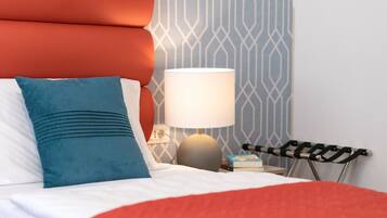 A neatly made bed with a red blanket, a blue pillow, and a white pillow. There is a lamp with a white shade on a bedside table. A book and a small box are also on the table.