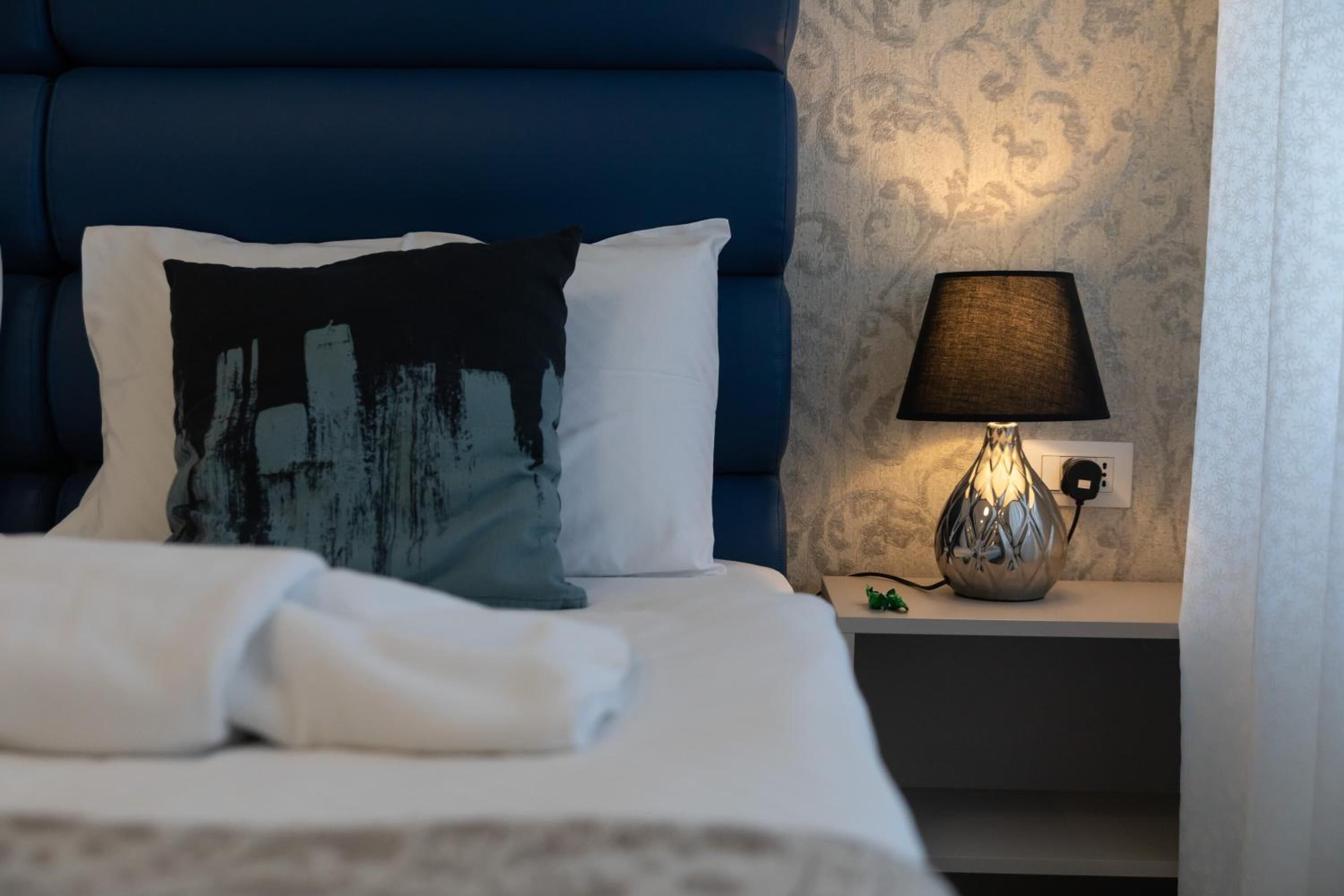 A neatly made bed with white linens, a blue headboard, and a decorative pillow. There is a bedside lamp on a nightstand beside the bed.