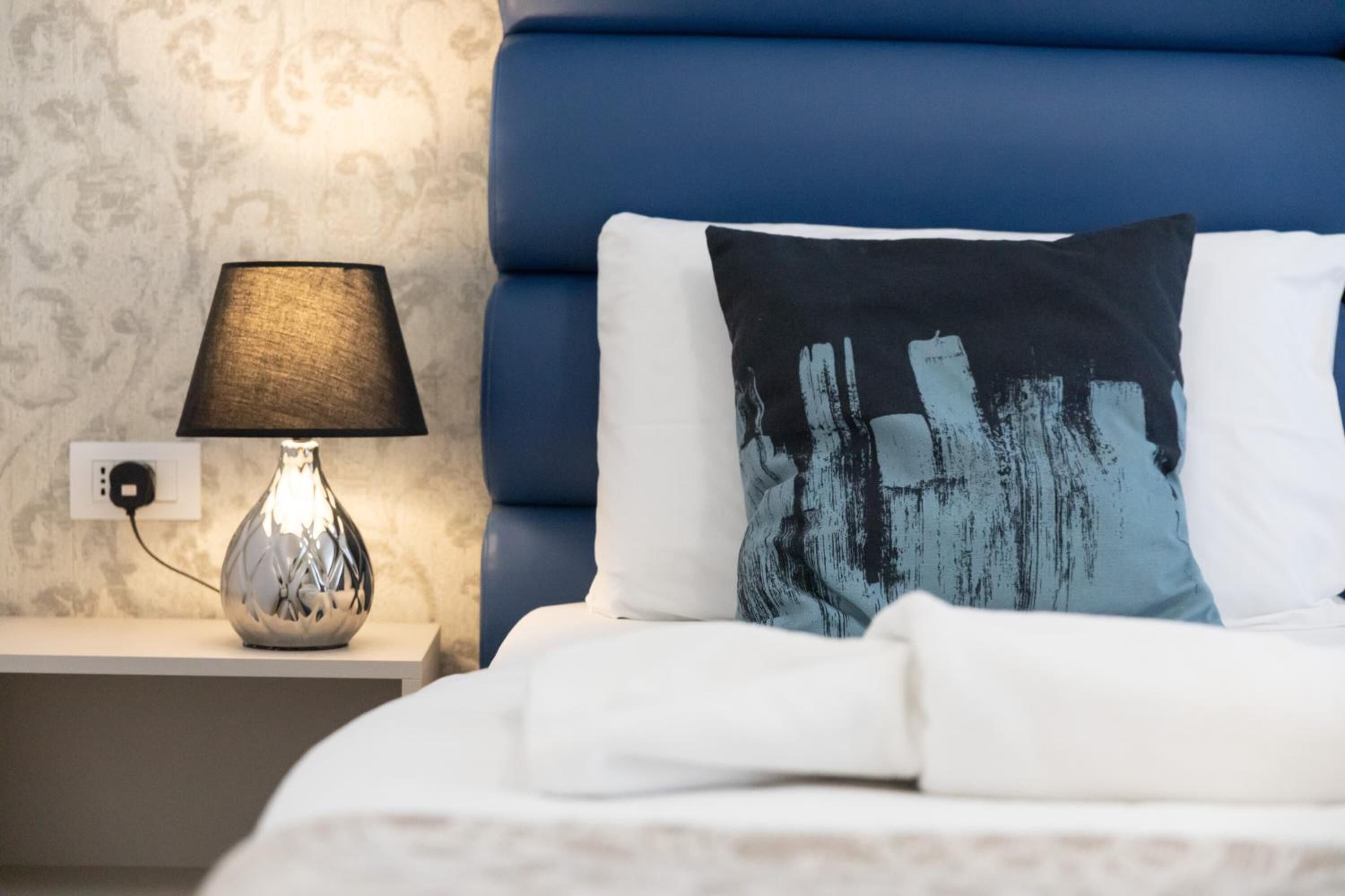 Ein ordentlich bezogenes Bett mit weißer Bettwäsche, ein blaues Kissen mit abstraktem Muster und eine Nachttischlampe mit strukturierter Lampenschirm auf einem weißen Nachttisch.