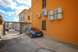 Uma rua estreita com um carro azul estacionado na lateral, um edifício com várias unidades de ar condicionado na parede e uma placa de estacionamento.