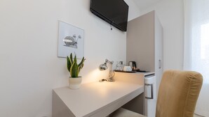 Una habitación moderna con un escritorio blanco, una planta en maceta, una lámpara, un televisor de pantalla plana y una silla color beige.