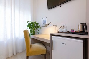 Habitación de hotel compacta con escritorio, una silla amarilla, un refrigerador pequeño, una planta, un cuadro en la pared y un televisor montado sobre el escritorio.