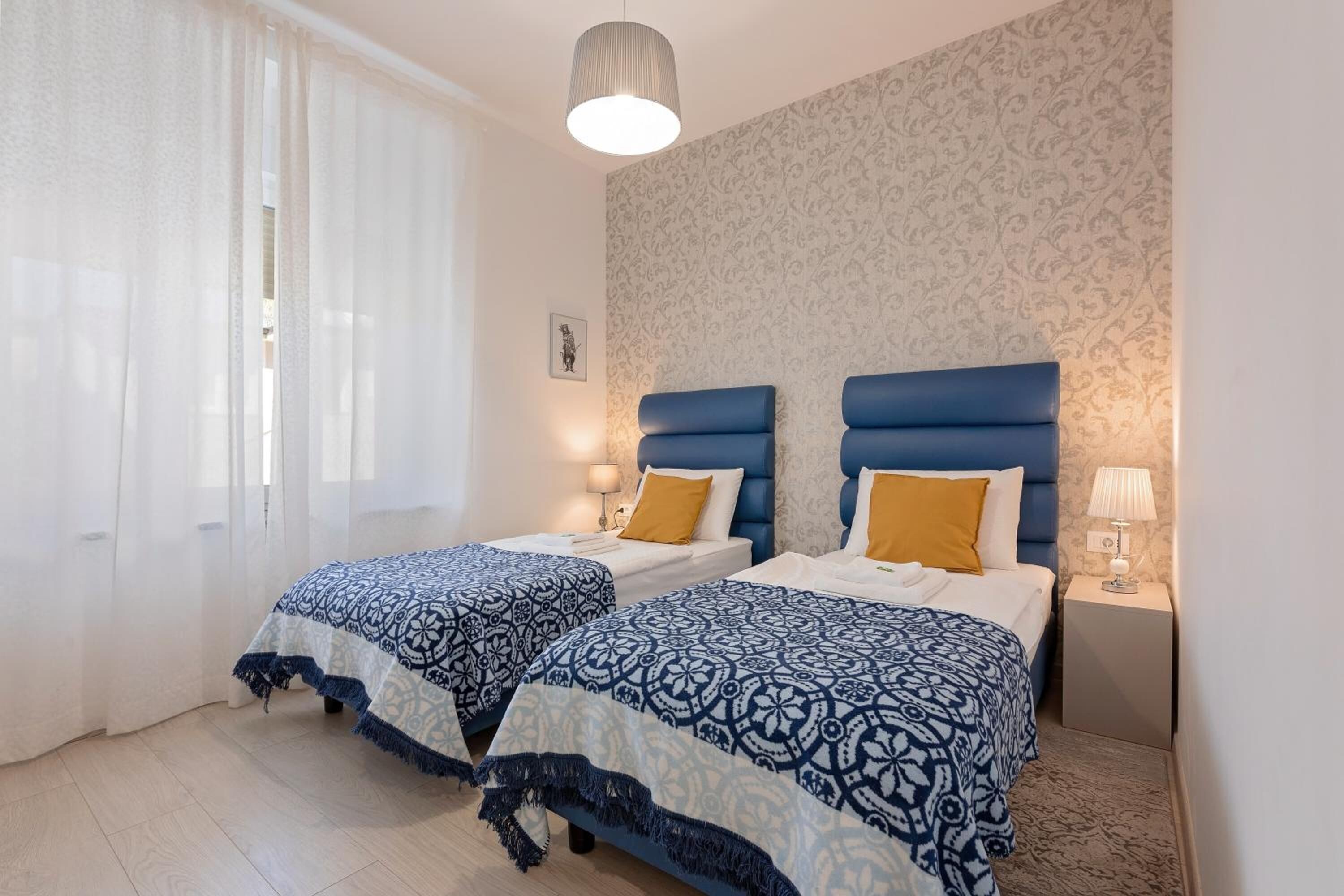 A hotel room with two beds, each with a blue patterned bedspread and a white pillow. There is a small bedside table with a lamp on each side of the beds. The room has a patterned wallpaper and a light-colored floor.