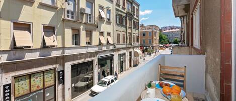 Una calle estrecha flanqueada por edificios, un balcón con una mesa preparada para el desayuno, y una furgoneta blanca estacionada en la vía.