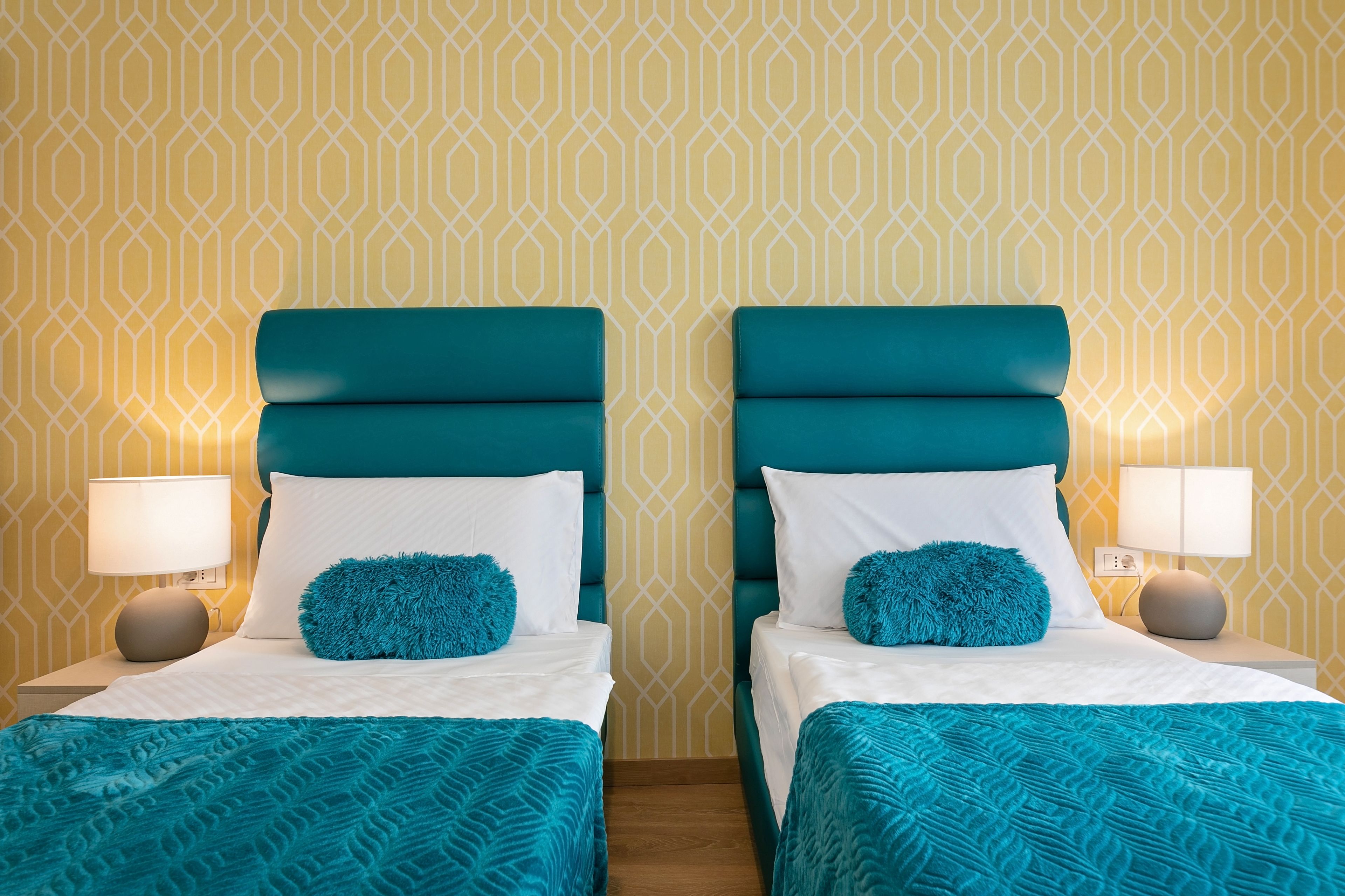 Ett hotellrum med två sängar, var och en med turkosa sänggavlar och vita sängkläder. Det finns matchande turkosa prydnadskuddar och en turkos pläd på varje säng. Rummet har en tapet med geometriskt mönster och två sänglampor.