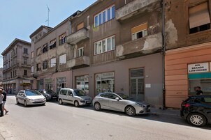 Ένας δρόμος με σταθμευμένα αυτοκίνητα και ένα κτίριο με μια πινακίδα που αναγράφει: «ΓΝΩΣΤΕΣ ΜΑΡΚΕΣ, ΧΑΜΗΛΕΣ ΤΙΜΕΣ».