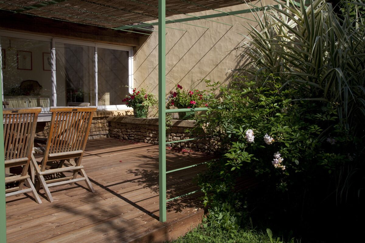 Una terraza de madera con sillas, una pérgola y un jardín con plantas en flor.