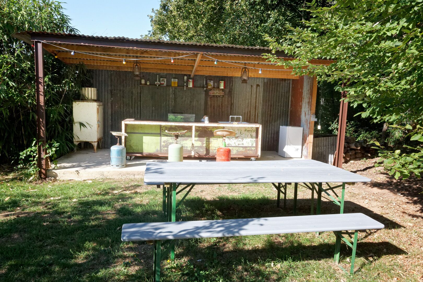 An outdoor patio area with a table and benches, surrounded by a rustic wooden structure with a corrugated metal roof.