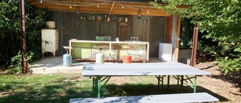 An outdoor patio area with a table and benches, surrounded by a rustic wooden structure with a corrugated metal roof.