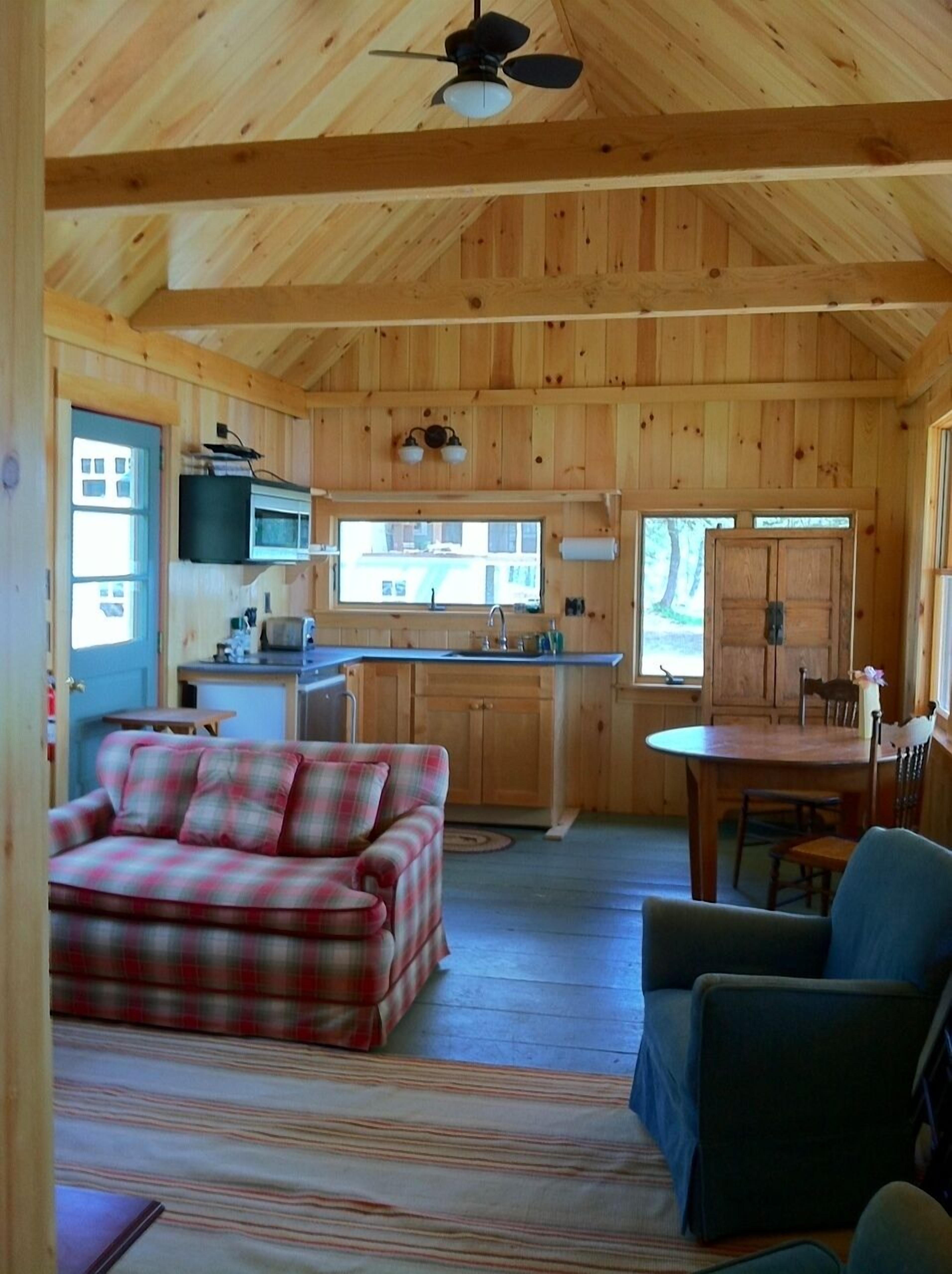 Un interior de cabaña acogedor con un sofá de cuadros, una mesa de comedor y una zona de cocina con microondas y fregadero.