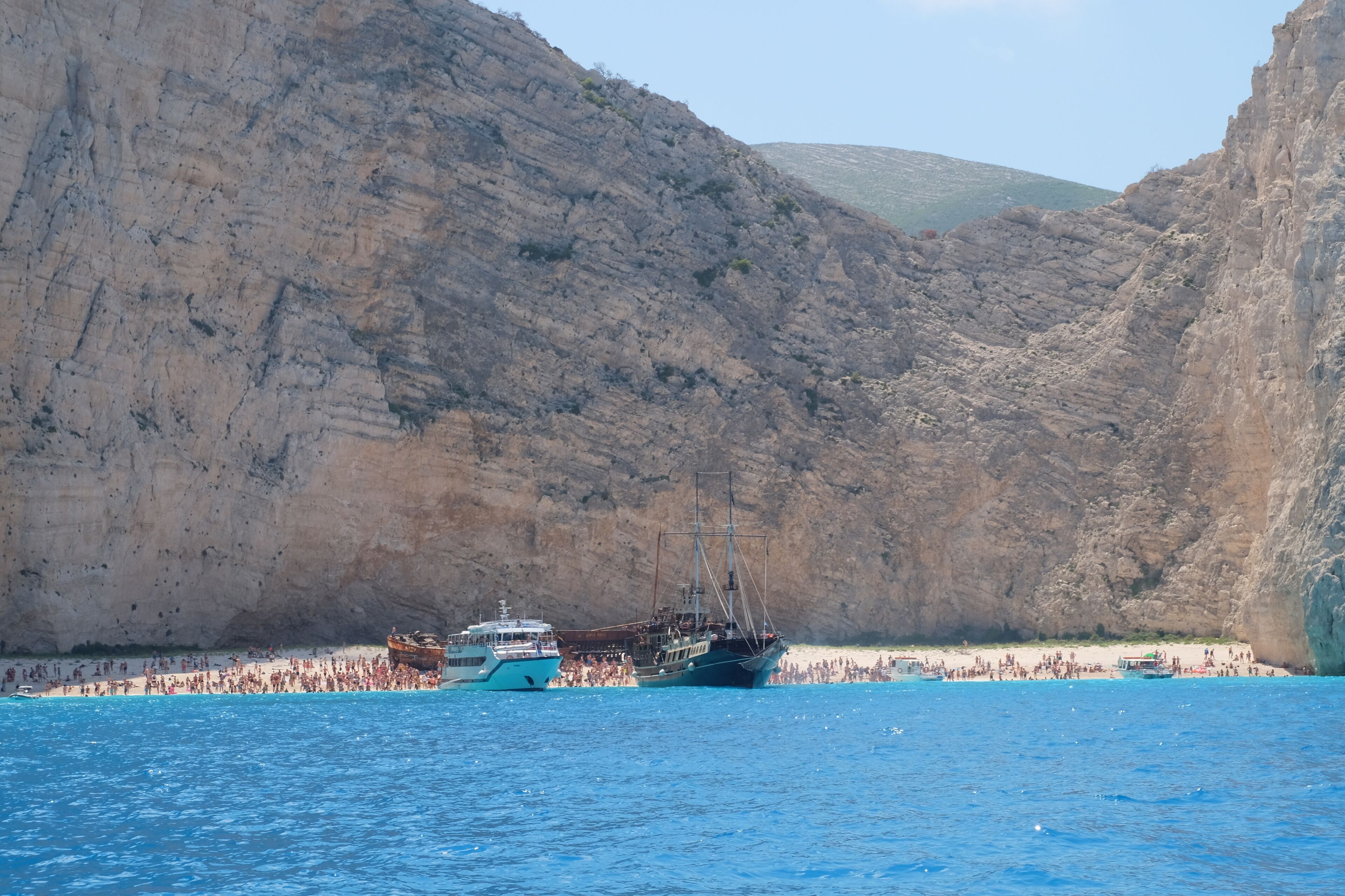Kustlandschap met een groot schip afgemeerd bij een rotsachtige klif, een strand met mensen en helderblauw water.