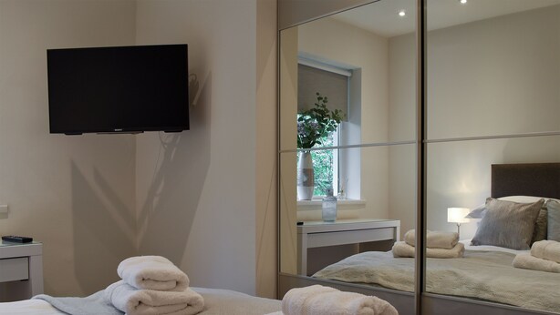 Ein modernes Hotelzimmer mit einem Bett, einem an der Wand montierten Flachbildfernseher, einem Nachttisch mit Lampe und einem großen Spiegel, der das Zimmer reflektiert.