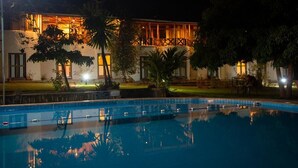 Vista nocturna de un hotel con piscina, un edificio bien iluminado y vegetación circundante.