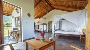 Una habitación con techo de madera, cama con dosel blanco, una mesa de madera con un jarrón de flores y vistas al exterior por una puerta abierta.