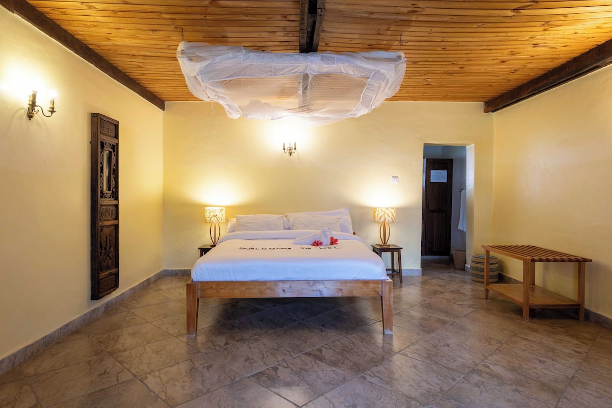 Quarto com cama de madeira, duas luminárias de cabeceira, um banco de madeira e um lustre.