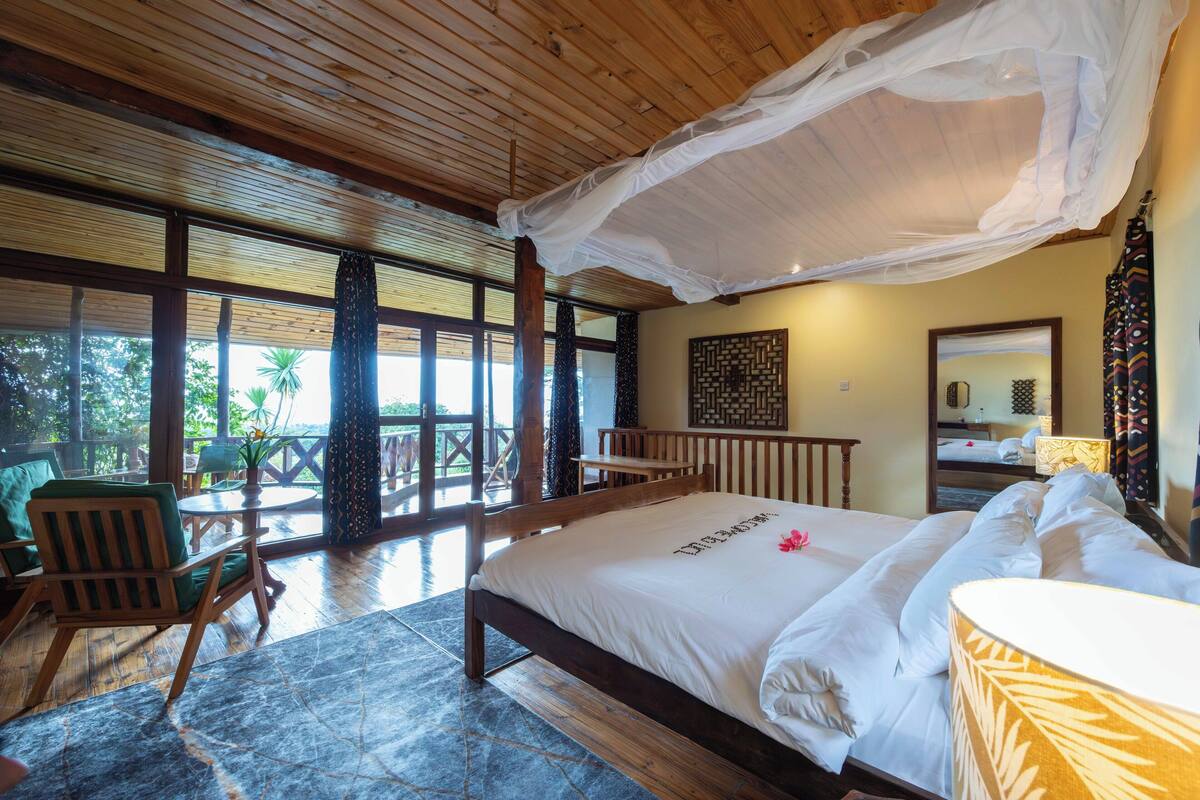 Quarto com cama de madeira, dossel, vista para o exterior, uma cadeira verde e piso de madeira.