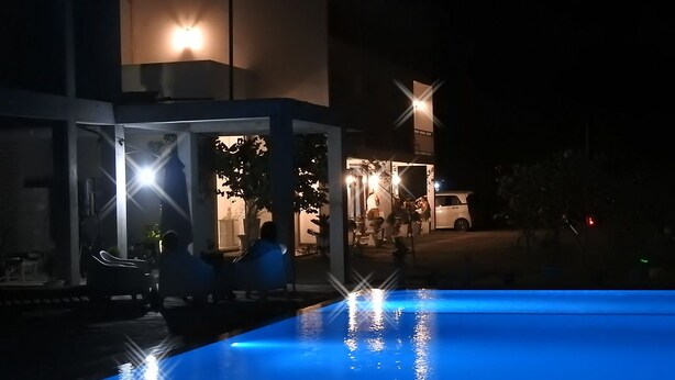 Vista nocturna de un área de piscina con una fuente iluminada en azul, una zona de estar cubierta y un edificio con ventanas iluminadas.