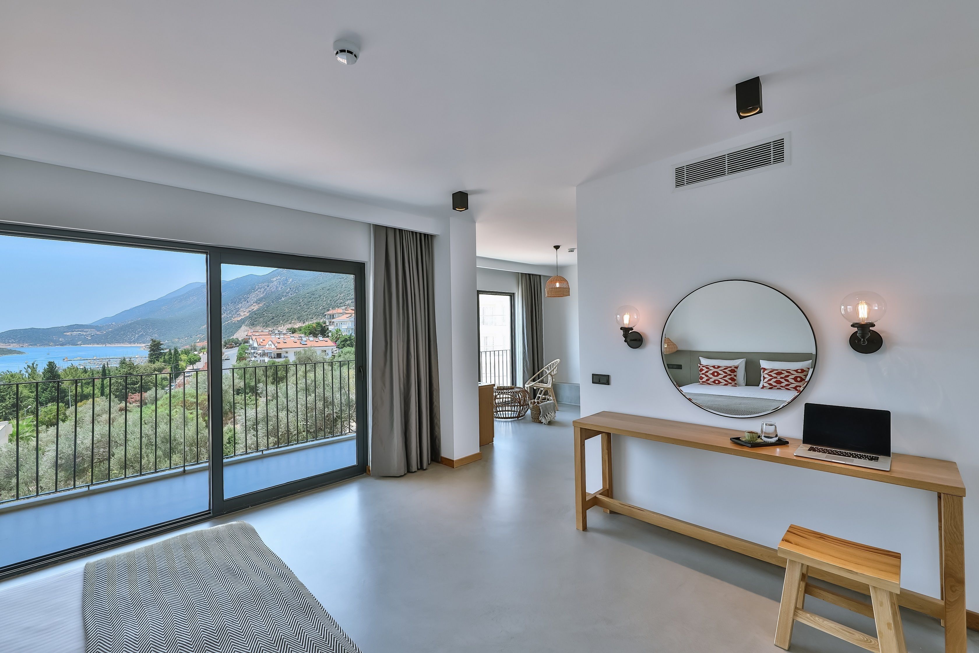 Una habitación de hotel moderna con un balcón grande, un escritorio de madera, una computadora portátil y un espejo redondo.