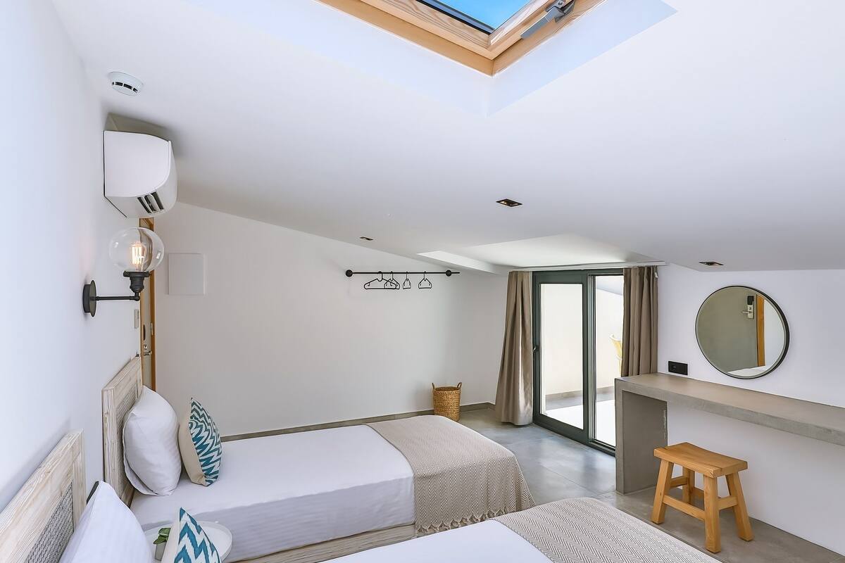 Una habitación de hotel moderna con tragaluz, dos camas, un taburete pequeño, un tocador con espejo y vistas al exterior.